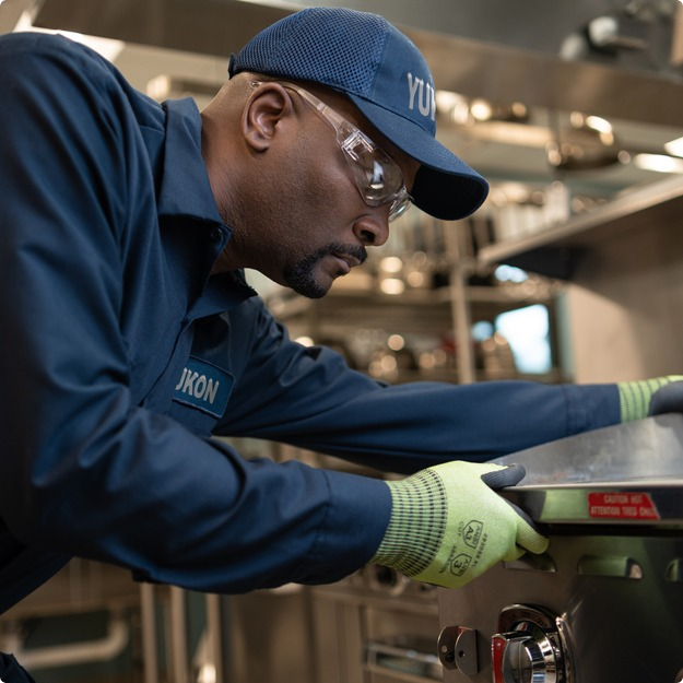 Technician in a cap and safety glasses servicing a commercial range in a restaurant kitchen