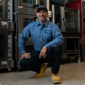 Smiling technician in a blue uniform and cap kneeling beside commercial ovens